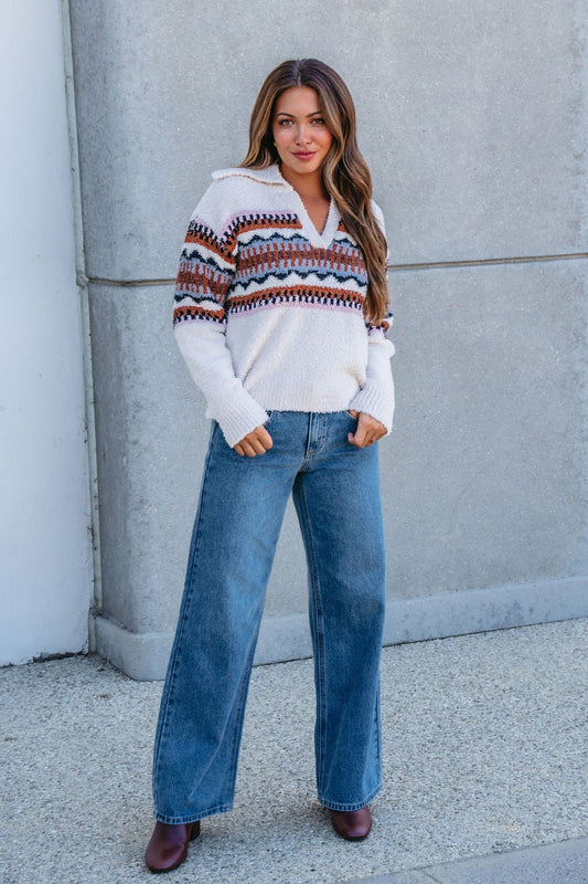 Woman wears the Thread and Supply Tan Aztec Bernese Sweater with wide-leg jeans and brown boots, hands in pockets by a gray wall.