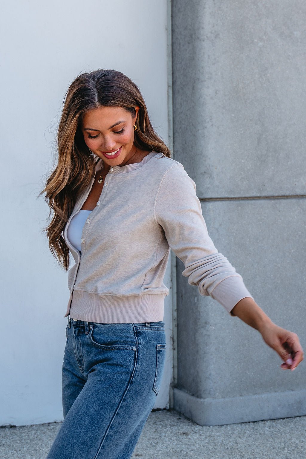 A woman smiles and looks down while walking outdoors in the Thread and Supply Taupe Monica Sweater Cardigan.