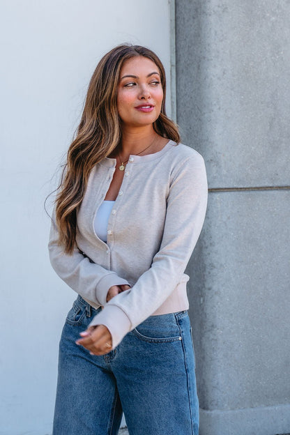 Woman with long brown hair smiles by a concrete wall in blue jeans and a Thread and Supply Taupe Monica Sweater Cardigan.