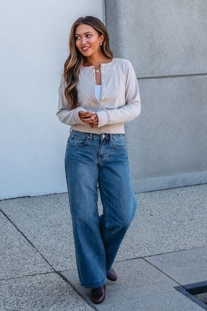 Woman with long hair in a Thread and Supply Taupe Monica Sweater Cardigan, white top, jeans, and brown boots stands on a sidewalk.
