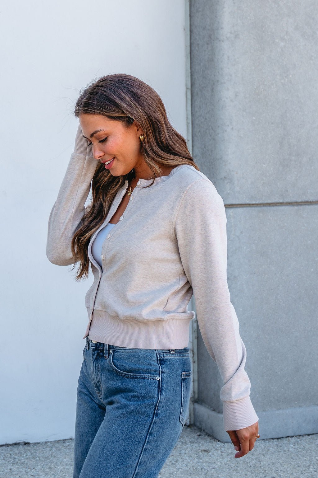 A woman smiles, touching her hair, in a Thread and Supply Taupe Monica Sweater Cardigan, white top, and jeans by a gray wall.
