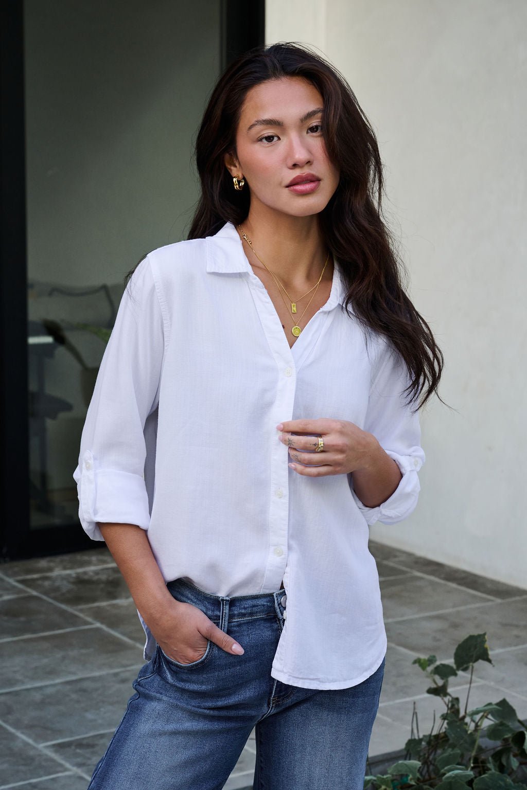 A woman with long dark hair wears the Thread and Supply White Button Down Presley Top and blue jeans outdoors, hand in pocket.