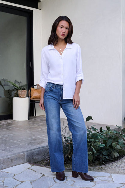 A woman stands outside in the Thread and Supply Presley white button down, blue jeans, brown shoes, with plants and a door behind her.