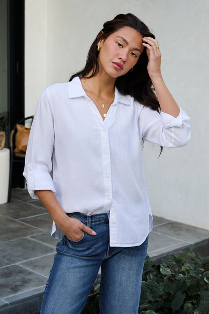 A woman wears the Thread and Supply White Button Down Presley Top with blue jeans, standing outside and touching her hair.
