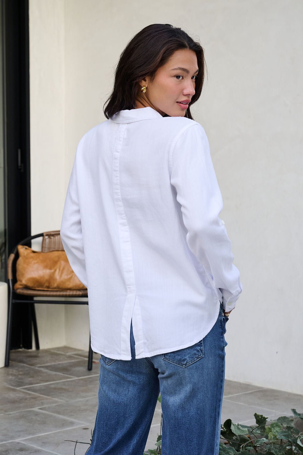 Woman with long brown hair wears the Thread and Supply White Button Down Presley Top and blue jeans outdoors, looking over her shoulder.