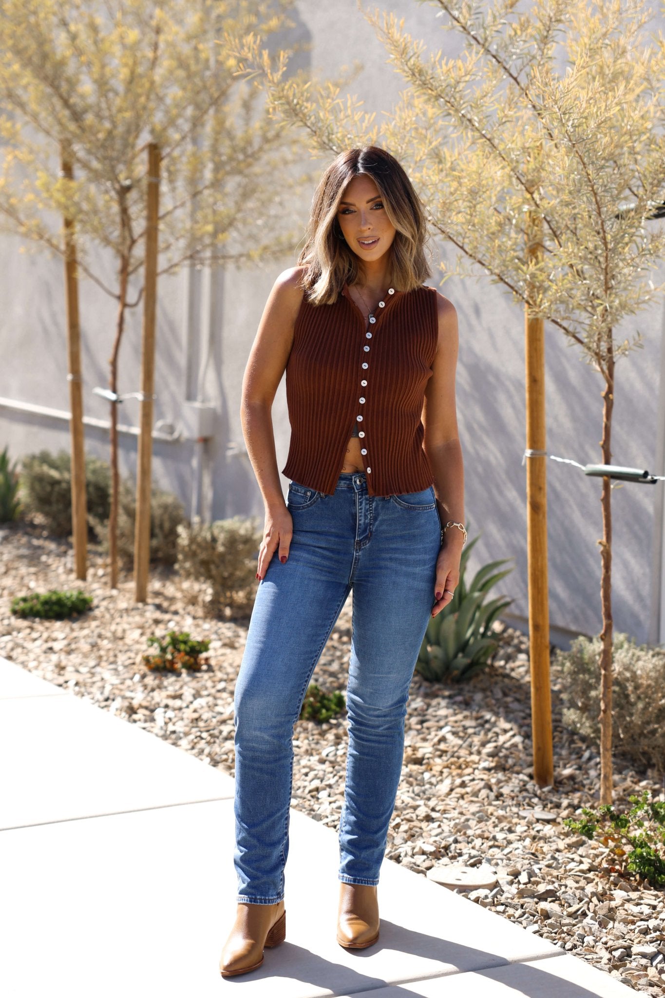 A woman in a brown sleeveless top and boots wears Thread & Supply Adrian Dark Wash Mid Rise Jeans outdoors near small trees.