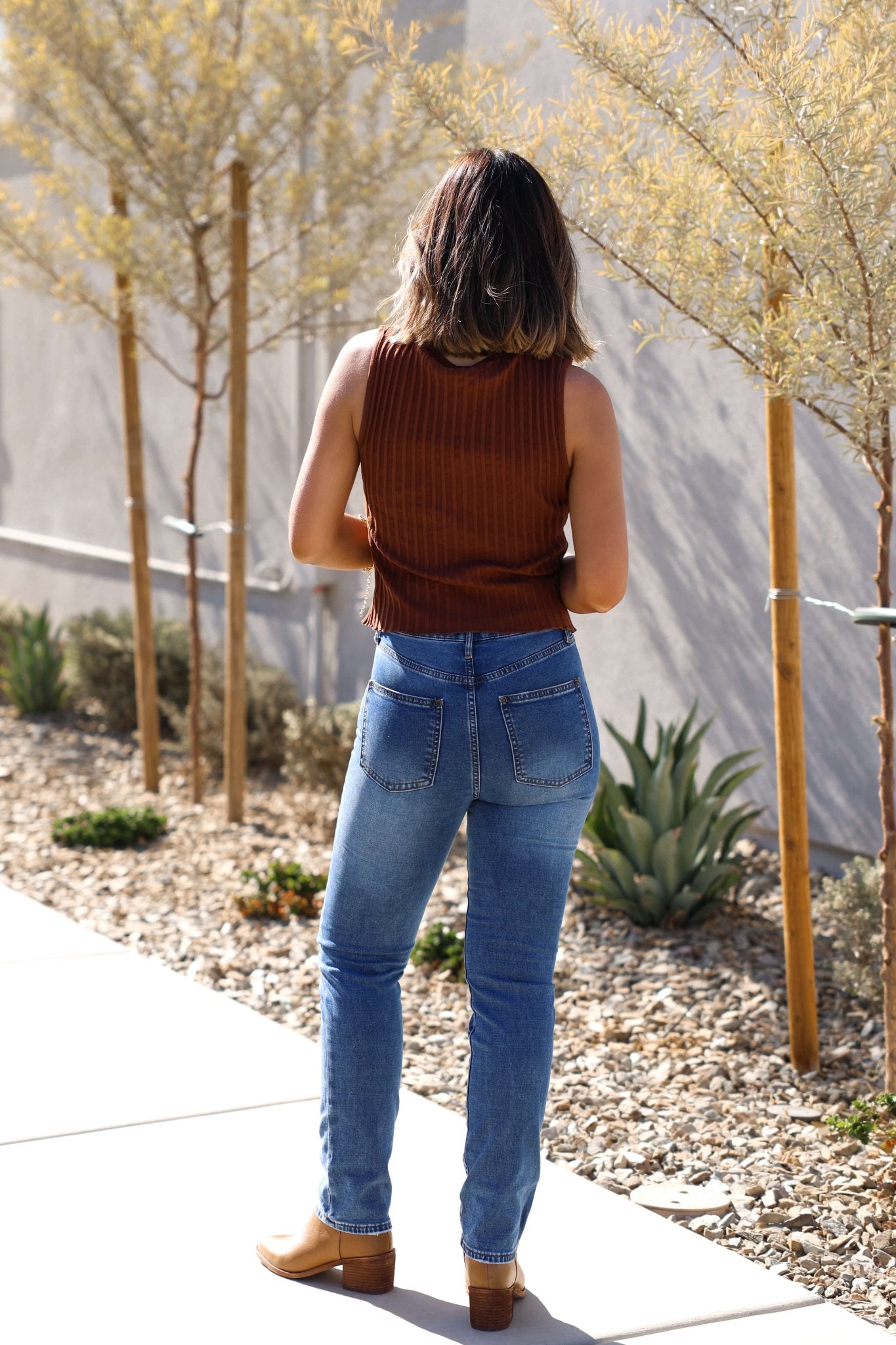 A woman in a brown sleeveless top and Thread & Supply Adrian Dark Wash Mid Rise Jeans stands on a sunny sidewalk near plants.