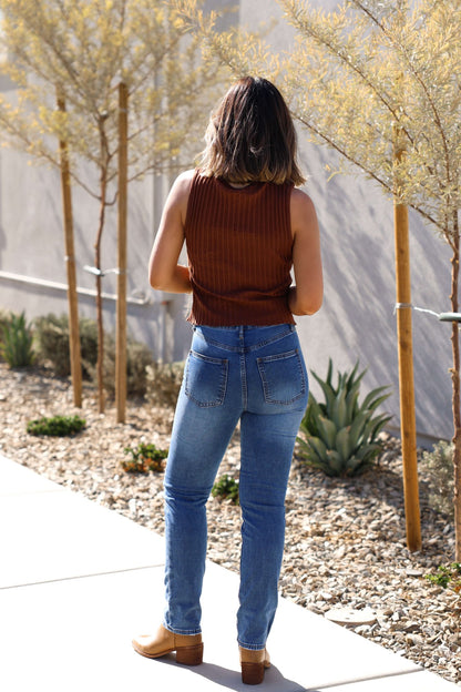 A woman in a brown sleeveless top and Thread & Supply Adrian Dark Wash Mid Rise Jeans stands on a sunny sidewalk near plants.