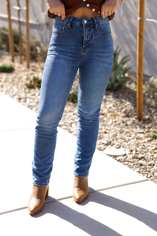 Person wears Thread & Supply Adrian Dark Wash Mid Rise Jeans, brown ankle boots, and a brown top on a sidewalk with plants and rocks.