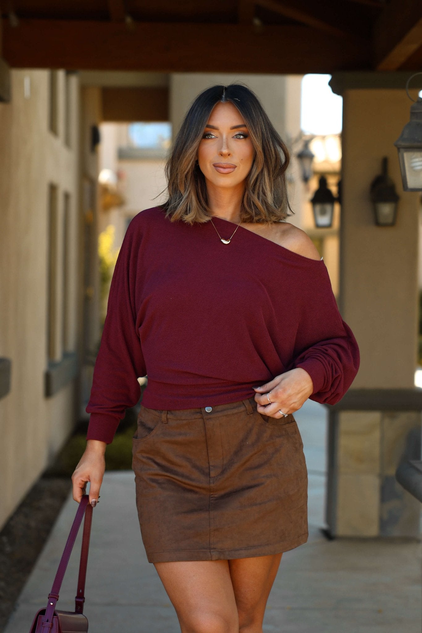 A woman in a burgundy off-shoulder top and Thread & Supply Brown Faux Suede Mini Skirt stands outdoors with a bag, lanterns, and buildings.