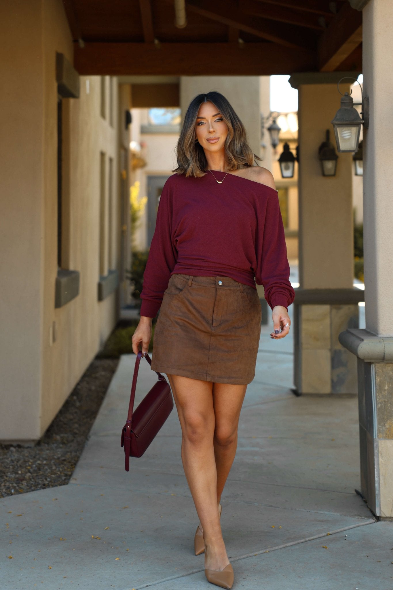 Woman in a Wine Off The Shoulder Crepe Top and brown skirt walks outside with a maroon handbag and beige heels.