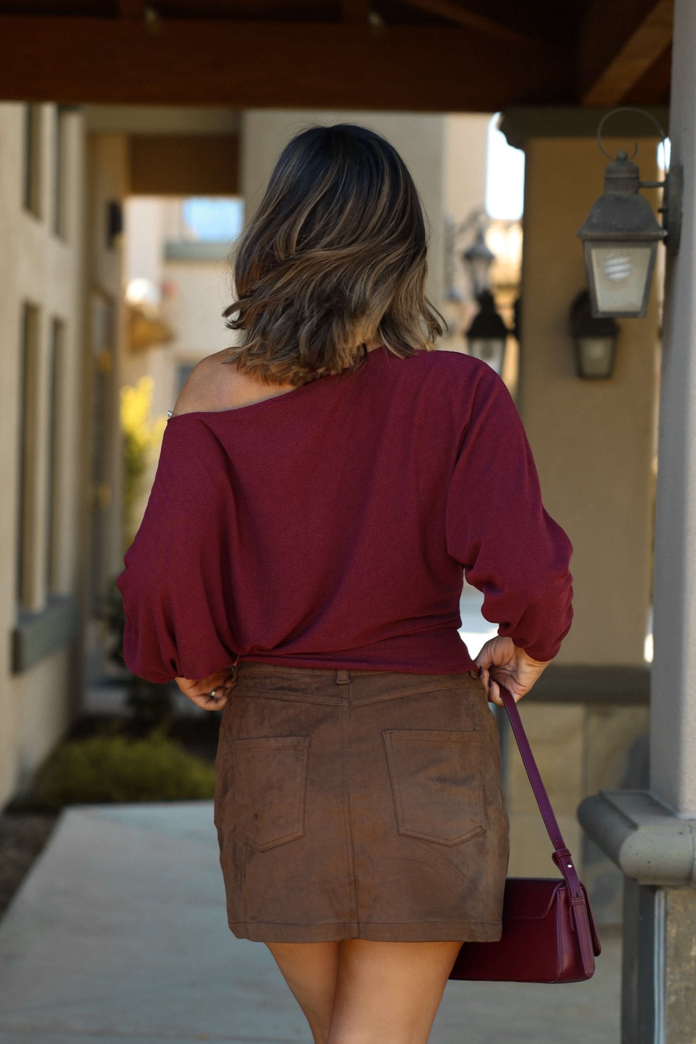 A woman with shoulder-length hair wears a maroon top and the Thread & Supply Brown Faux Suede Mini Skirt outdoors, back to camera.