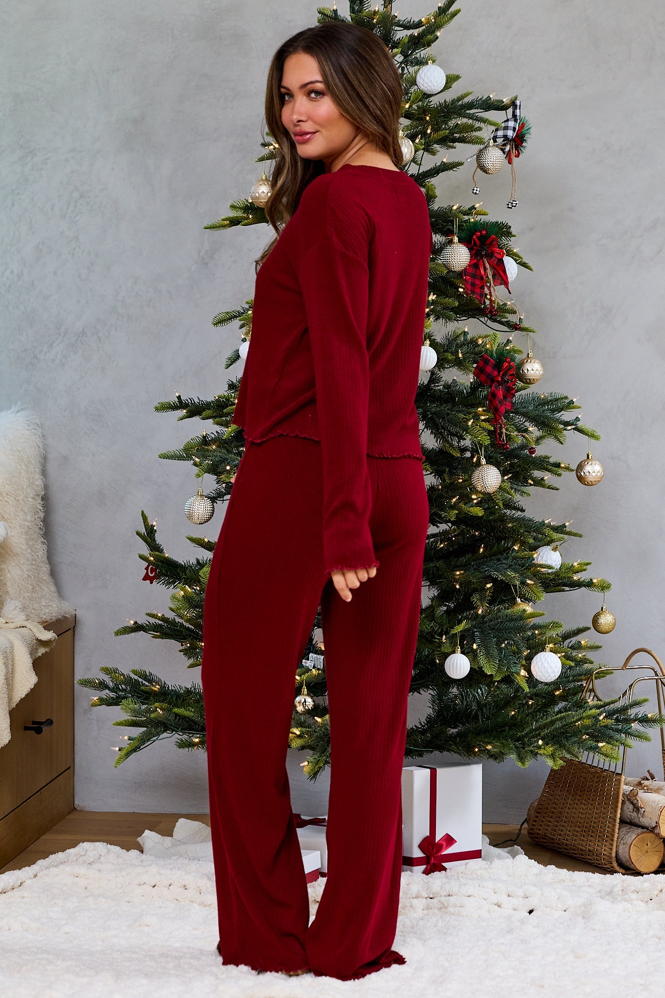 Woman in a Thread & Supply Burgundy Pajama Set stands by a Christmas tree with wrapped gifts beneath its glowing branches.