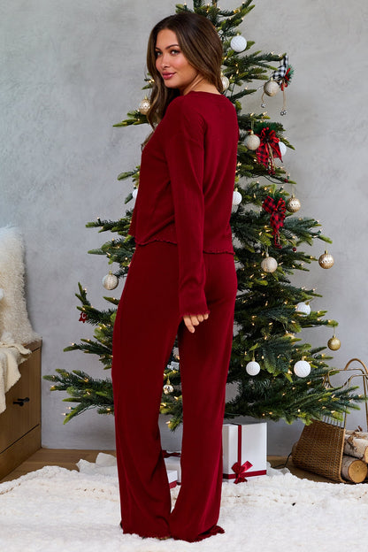 Woman in a Thread & Supply Burgundy Pajama Set stands by a Christmas tree with wrapped gifts beneath its glowing branches.