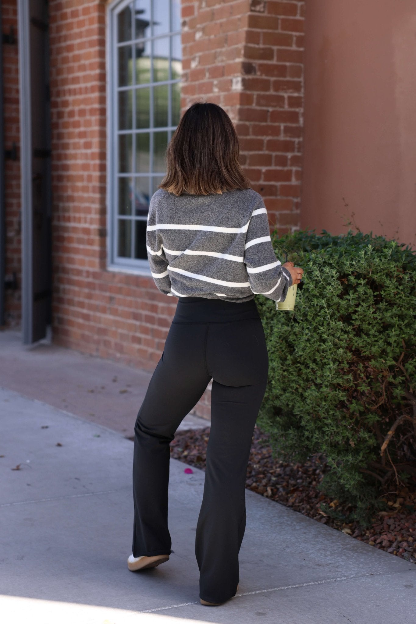A woman stands on a sidewalk in Thread & Supply Callie Black Flare Pants and a gray striped sweater, facing away.