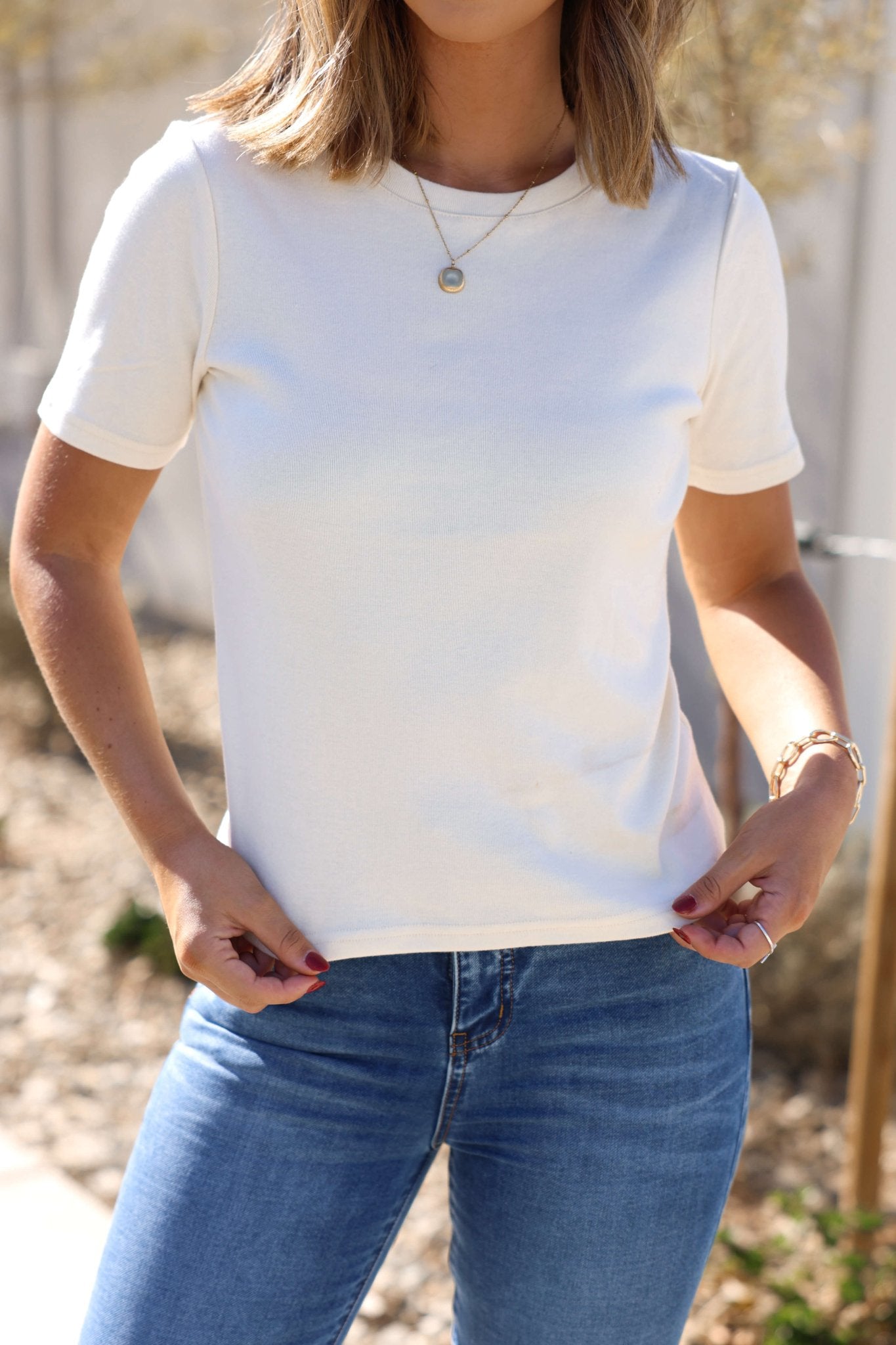 Woman wearing a Thread & Supply Sammie Cream Tee and blue jeans outdoors, holding the hem of her shirt.