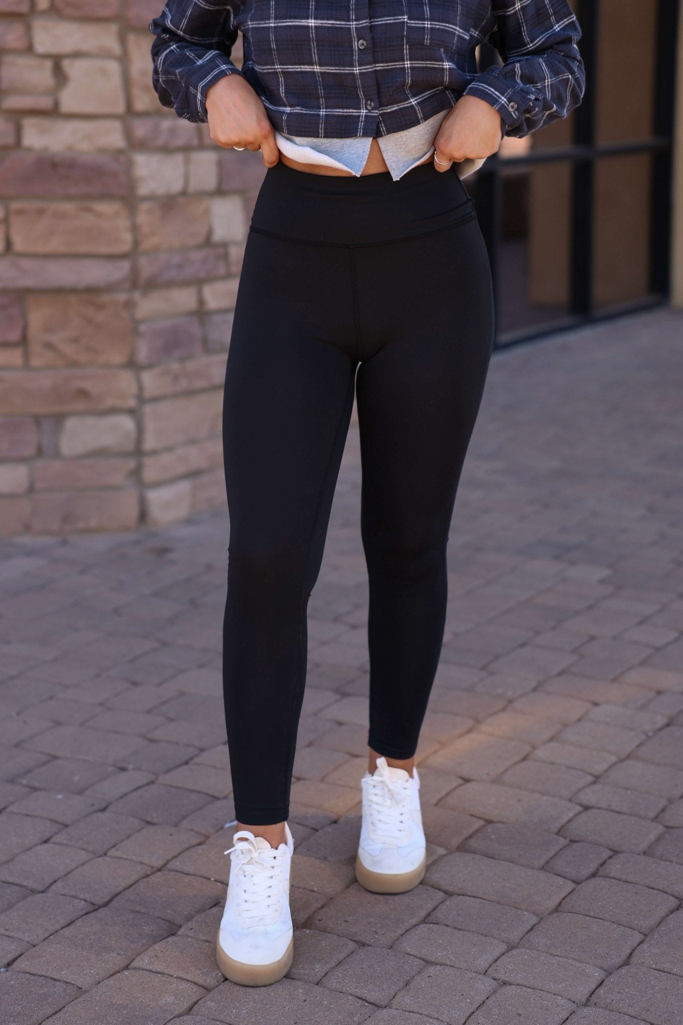 Wearing Thread & Supply Callie Black Leggings, a plaid shirt, and white sneakers, a person stands on a brick walkway adjusting their shirt.