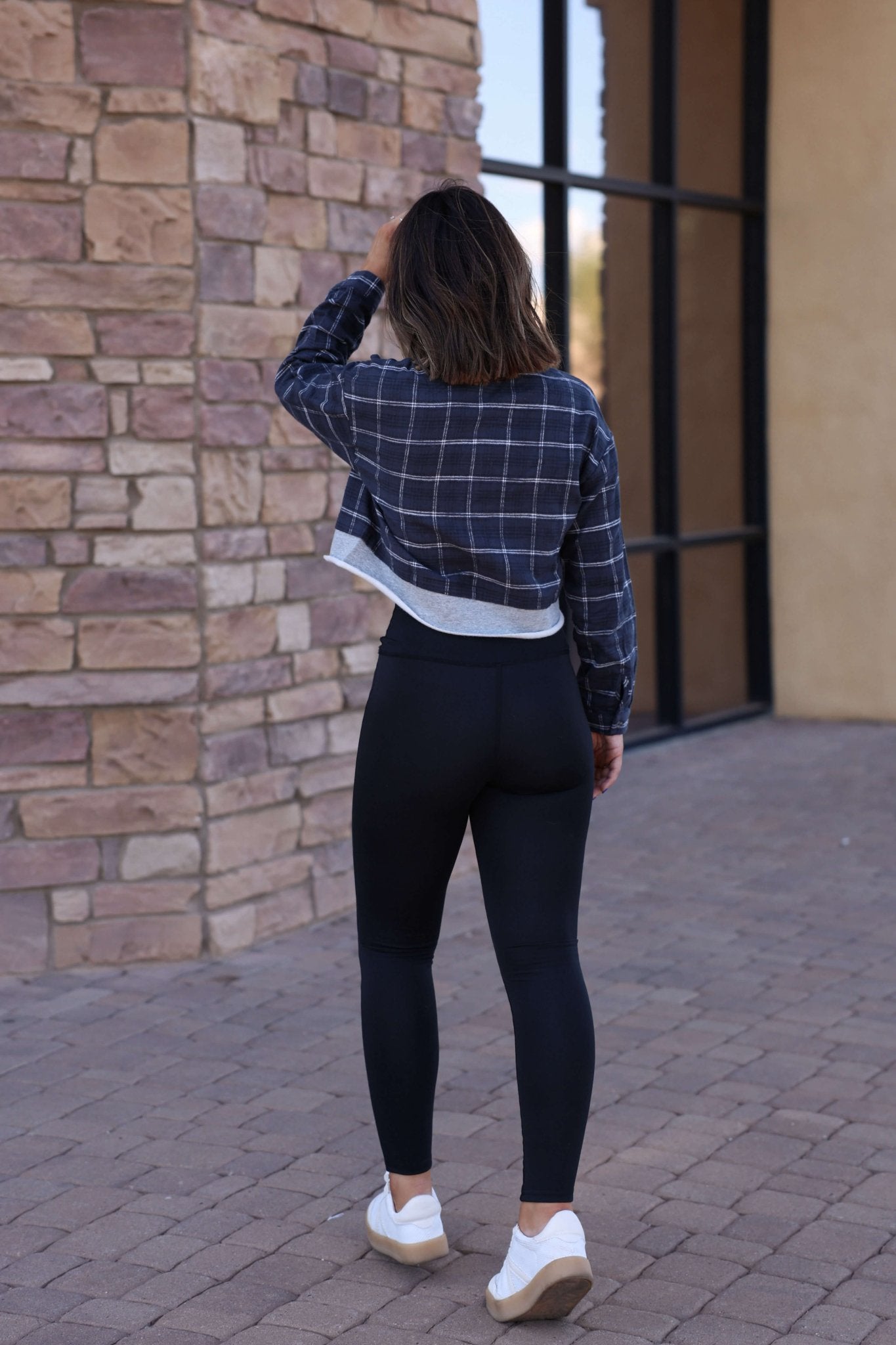 Wearing Thread & Supply Callie Black Leggings, white sneakers, and a plaid jacket, someone walks on a stone path beside a brick wall.