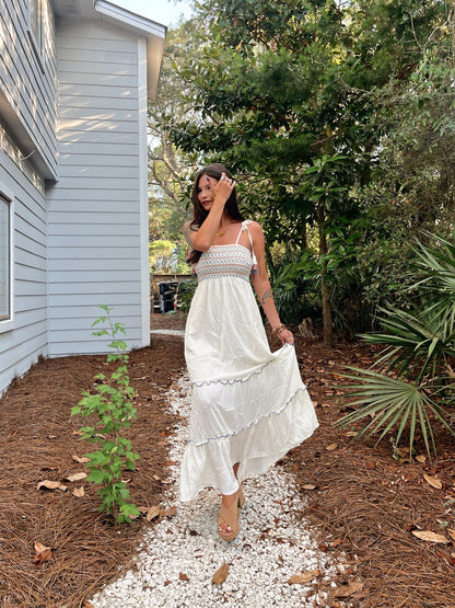 Woman in a Tie Strap Smocked Ivory Tiered Contrast Maxi stands on a gravel path by a light blue house, smiling and touching her hair.