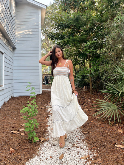 Woman in Tie Strap Smocked Ivory Tiered Contrast Maxi walks on a gravel path beside a light blue house and lush greenery.