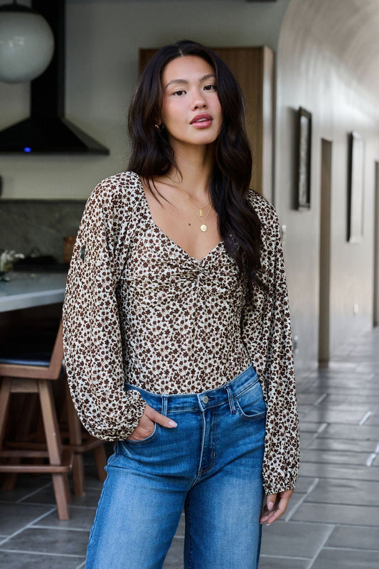 A woman wears the Touch Of Romance Brown Floral Bodysuit with blue jeans indoors, one hand in her pocket, looking at the camera.