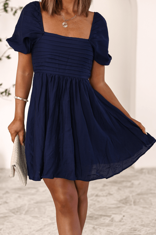 Woman wearing the Twilight Pleated Puff Sleeve Mini Dress, holding a light clutch on a textured floor.
