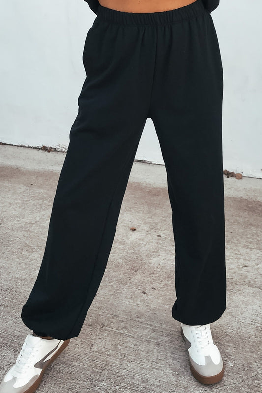 Person wearing Ultra Soft Brushed Fleece Sweatpants in black with white sneakers, standing on concrete against a light background.