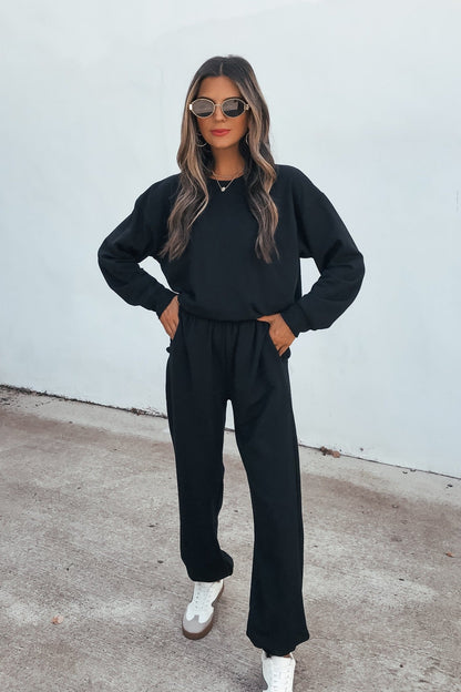 Woman in sunglasses wears the Ultra Soft Brushed Fleece Sweatshirt - Black with matching pants and sneakers, standing by a white wall.