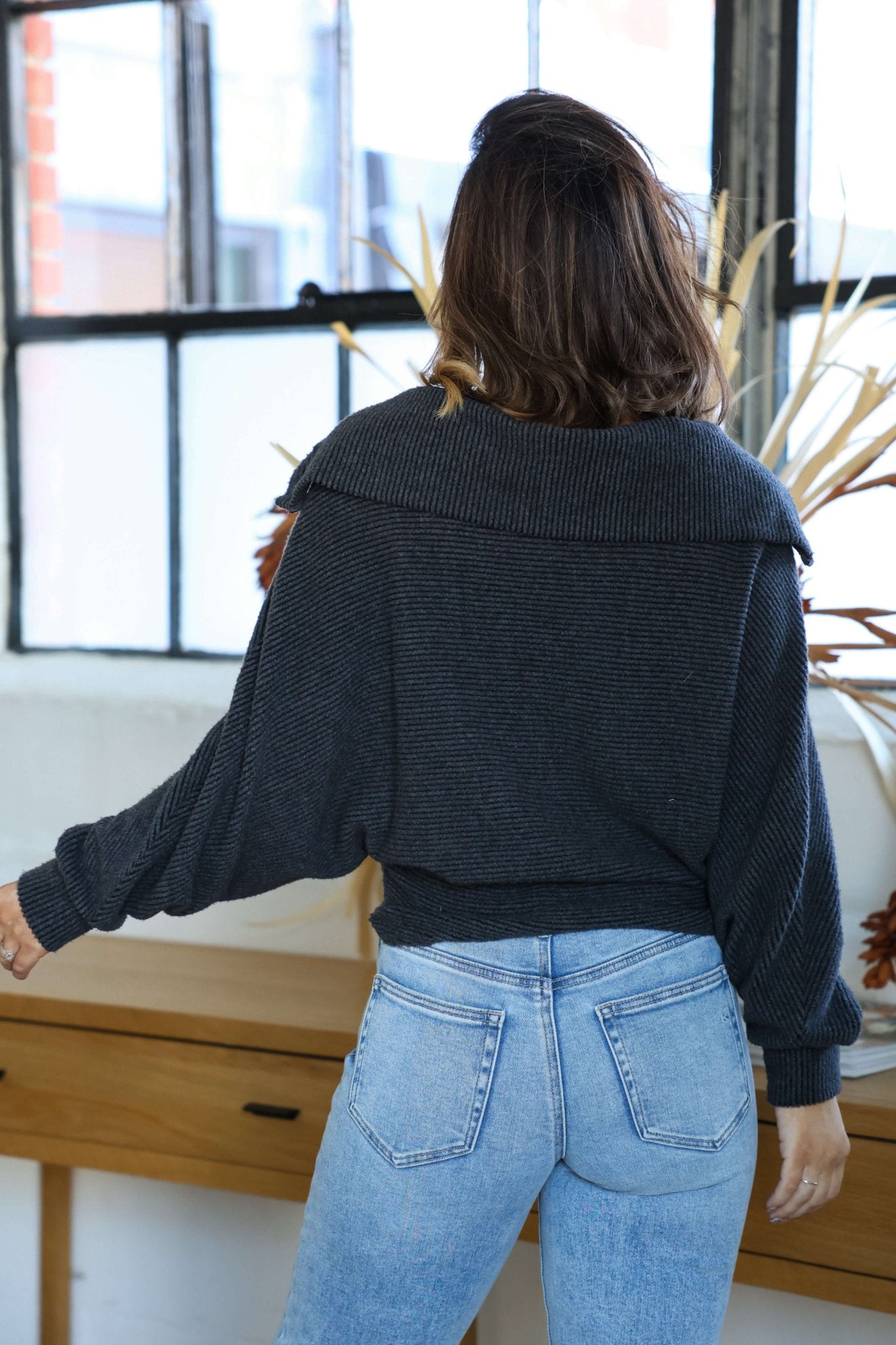 A woman in an Ultra Soft Charcoal Collared Pullover and blue jeans stands indoors, perfect for a cozy winter look.