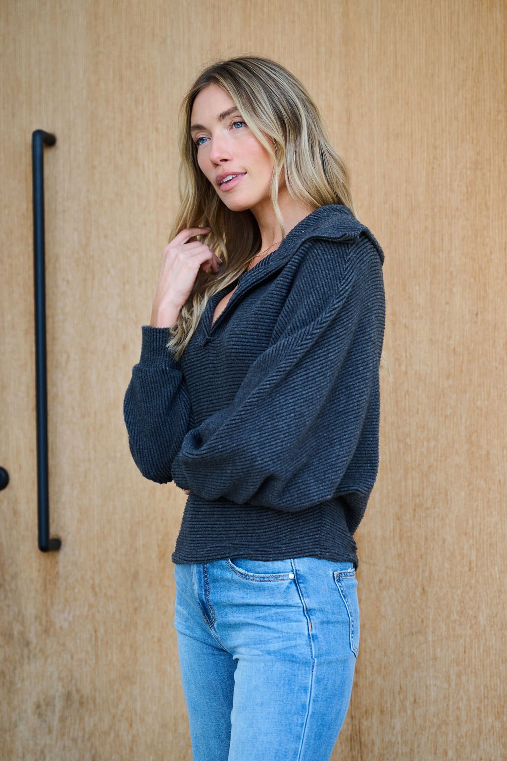 A woman with long blonde hair wears the Ultra Soft Charcoal Collared Pullover and light blue jeans, standing by a wooden wall.