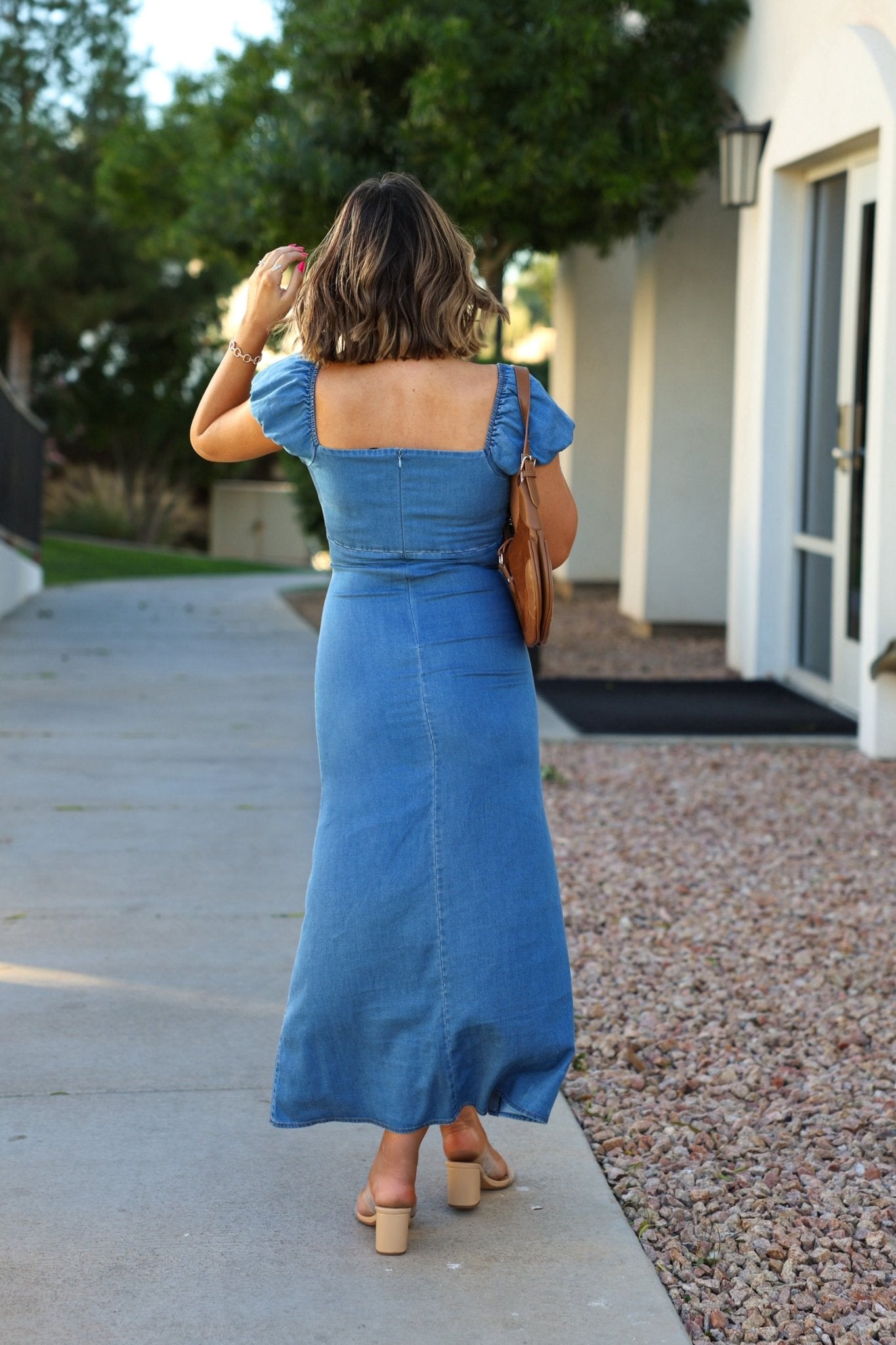 A woman in the Ultra Soft Denim Sweetheart Maxi Dress walks on a sidewalk near trees and a white building, carrying a brown handbag.