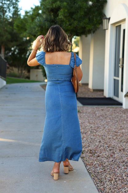A woman in the Ultra Soft Denim Sweetheart Maxi Dress walks on a sidewalk near trees and a white building, carrying a brown handbag.