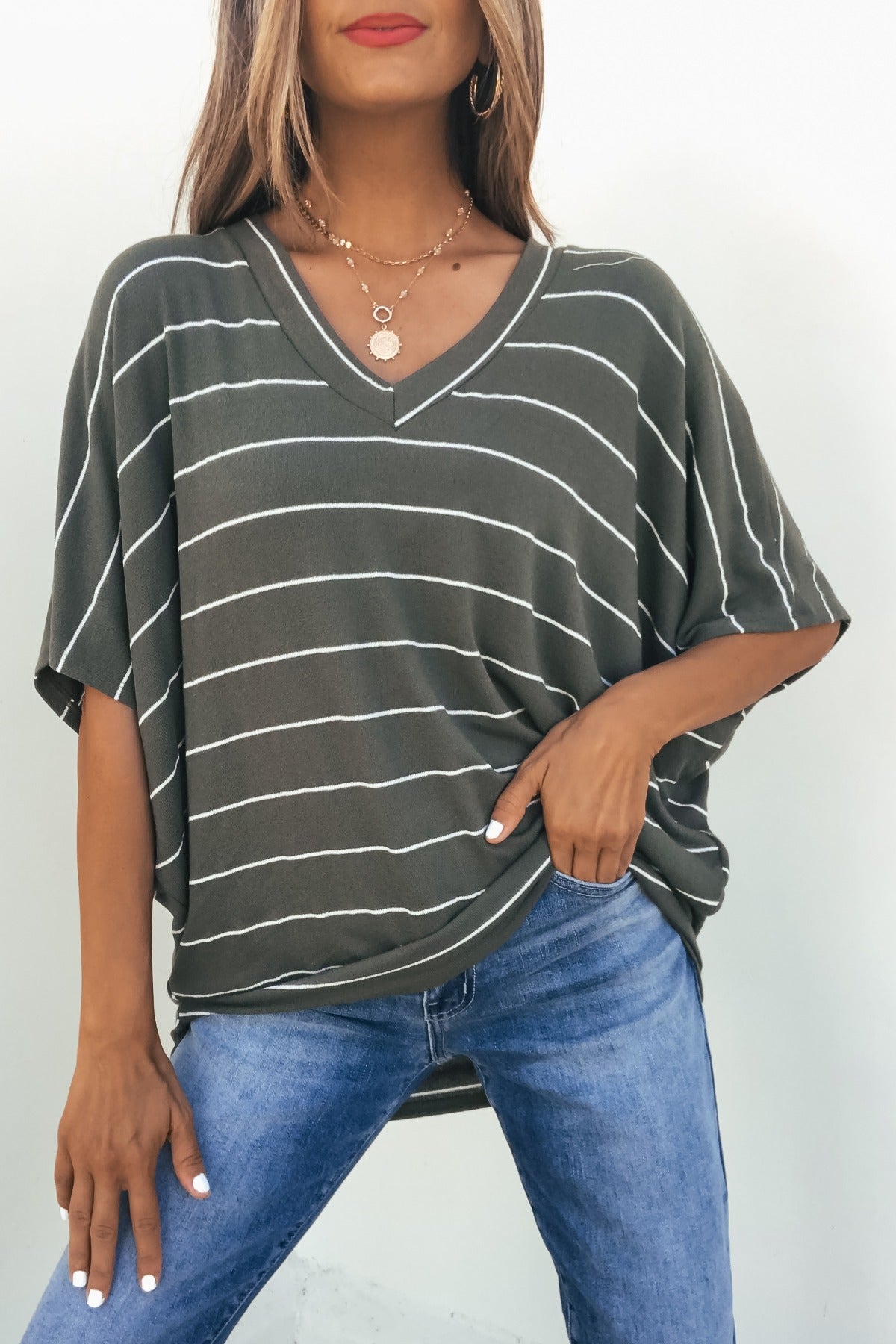 Woman wears the Unwind Stripe V Neck Top - Olive with blue jeans, one hand in pocket, standing by a plain wall.