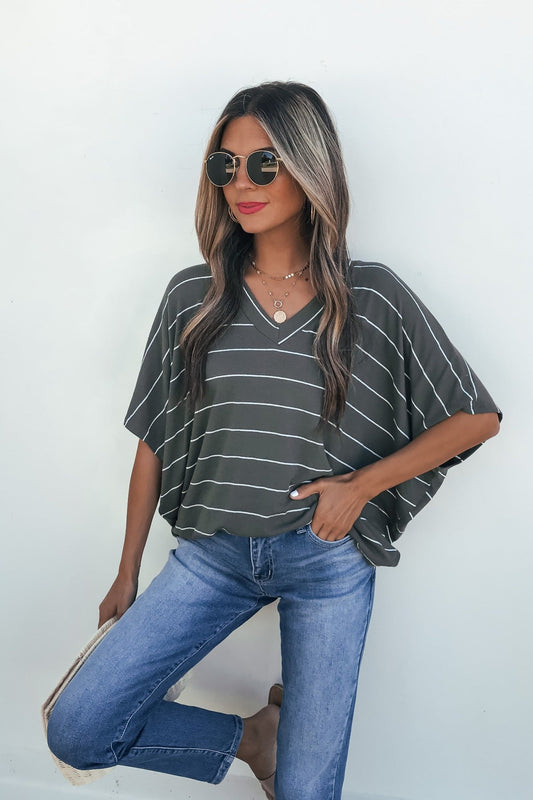 Woman in sunglasses wears the Unwind Stripe V Neck Top - Olive with blue jeans and holds a clutch against a white background.