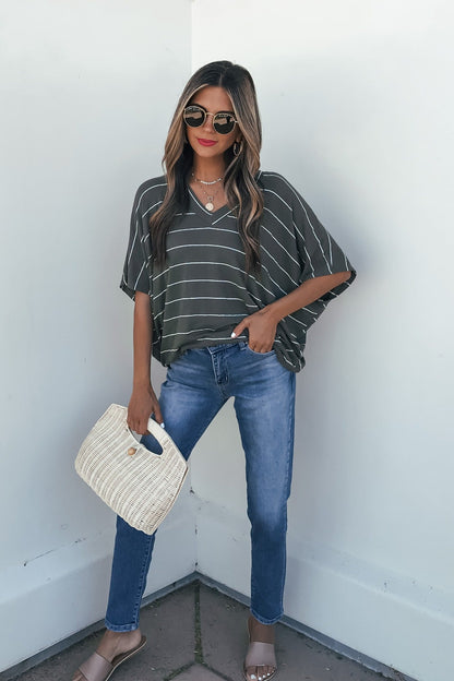 Woman in sunglasses, blue jeans, sandals, and Unwind Stripe V Neck Top - Olive stands with a woven bag against a white wall.