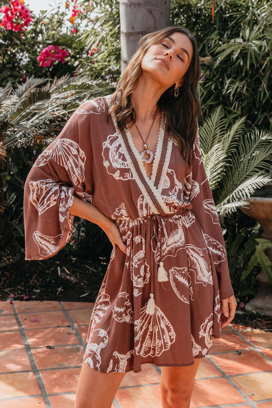 Woman in a Surf Gypsy Crochet Print Dress - Brown stands outdoors with lush greenery and blooming pink flowers in the background.