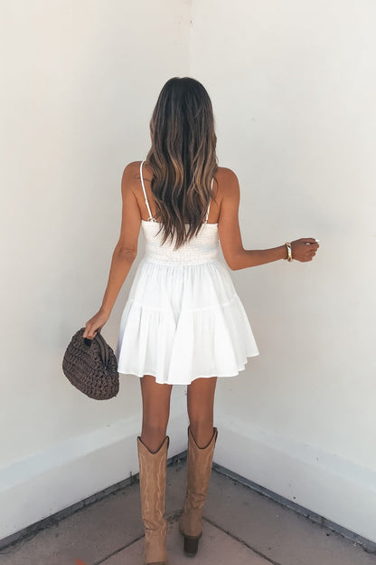 Woman with wavy hair wears the Valerie Embroidered Print Romper - White, tan boots, and holds a brown woven bag facing a white wall.