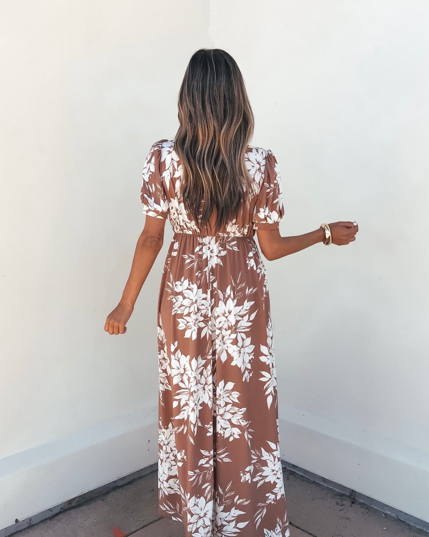Woman with long wavy hair models the Valerie Taupe V Neck Print Maxi Dress, facing a white wall with her back to the camera.