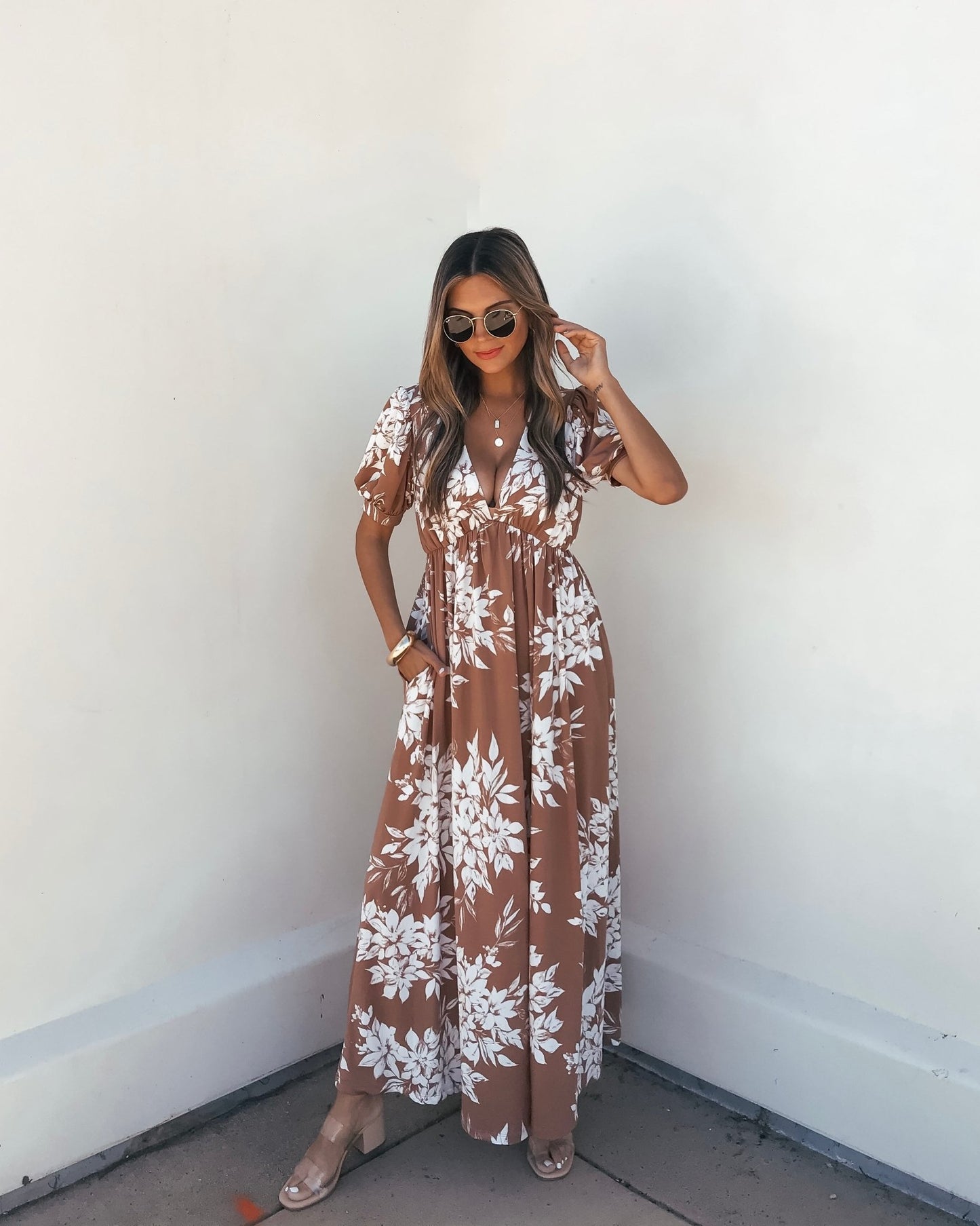 Woman wears the Valerie Taupe V Neck Print Maxi Dress, standing by a white wall and holding her hair.