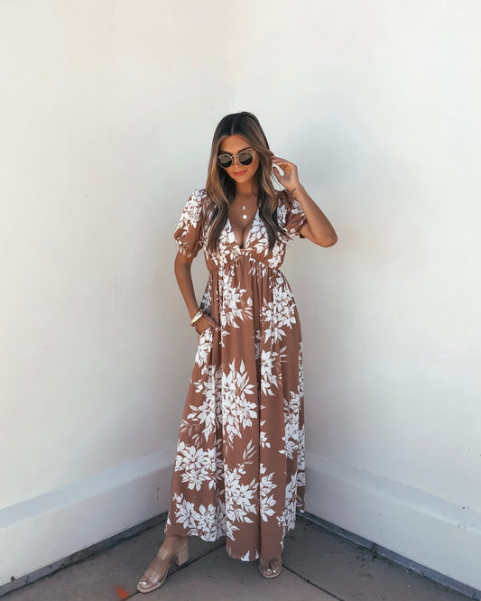 Woman wears the Valerie Taupe V Neck Print Maxi Dress, standing by a white wall and holding her hair.