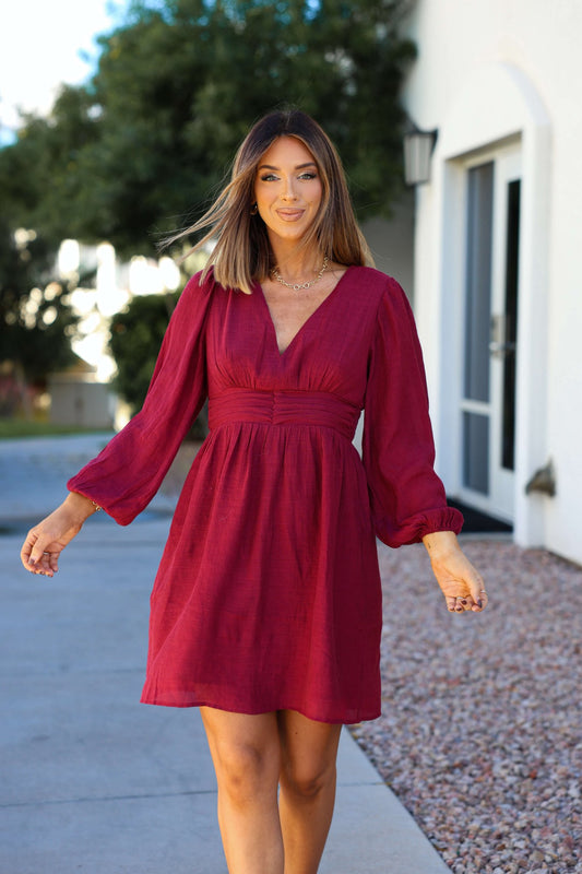 Woman with shoulder-length hair in the Vanessa Burgundy Ruched Mini Dress, walking outdoors on a sunny day near a white building.