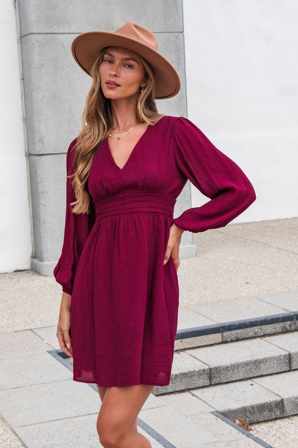 Model in the Vanessa Burgundy Ruched Mini Dress stands on stone steps outdoors, posing confidently with one hand on her hip.