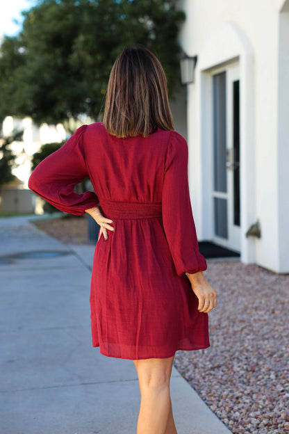 A woman with shoulder-length hair walks away, wearing the Vanessa Burgundy Ruched Mini Dress on the sidewalk.