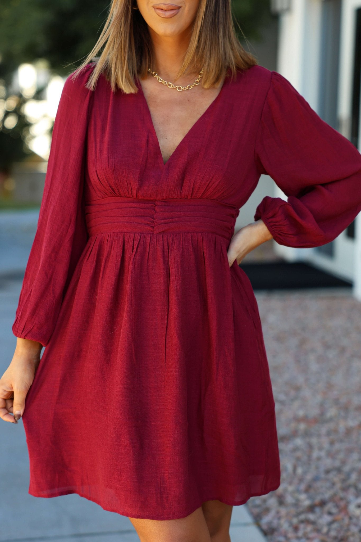 A woman in the Vanessa Burgundy Ruched Mini Dress with long sleeves and a V-neck stands outdoors, hand on hip.
