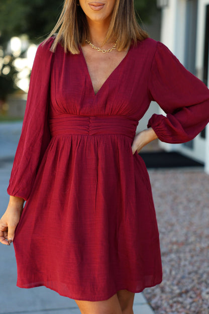 A woman in the Vanessa Burgundy Ruched Mini Dress with long sleeves and a V-neck stands outdoors, hand on hip.