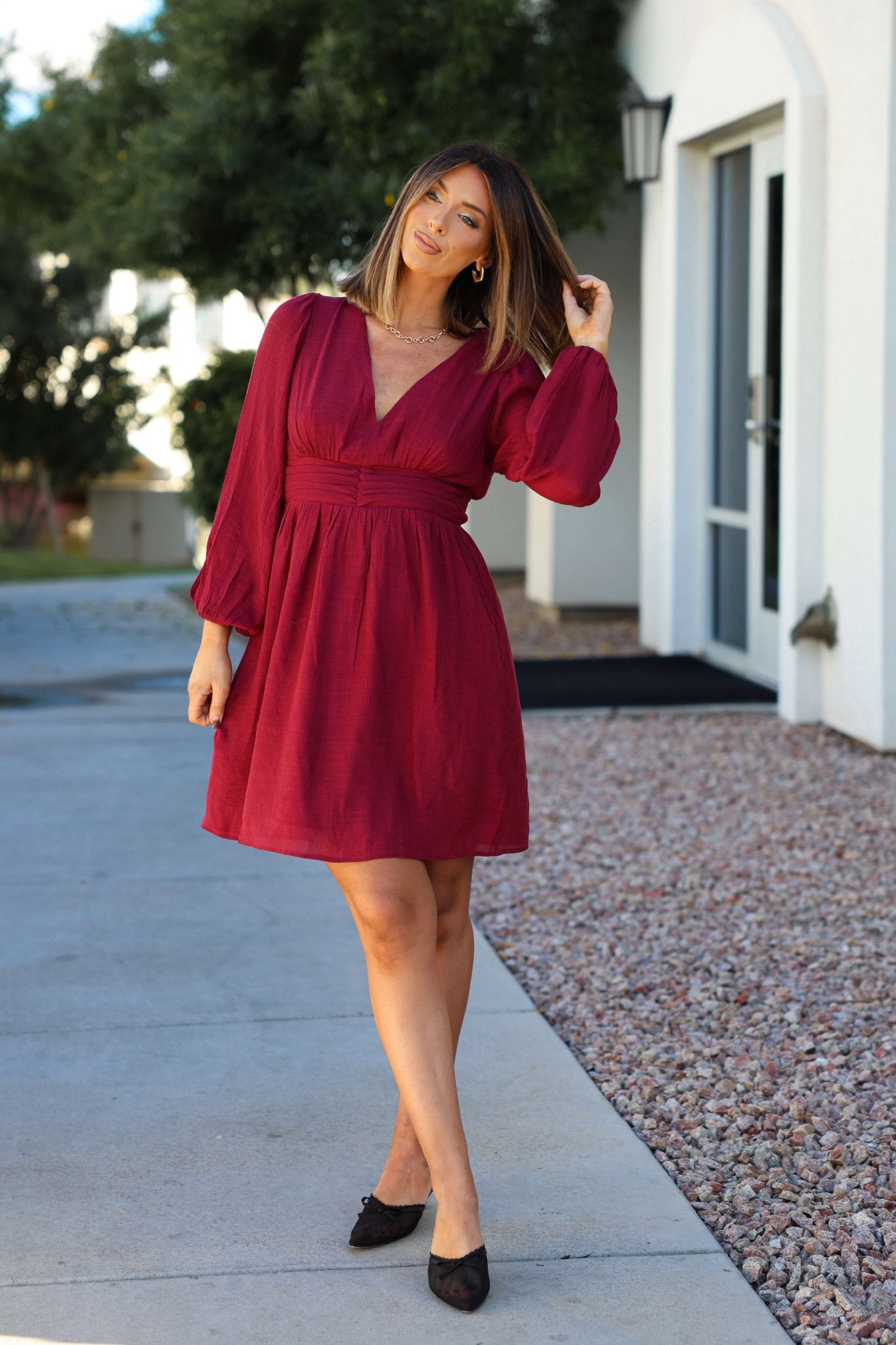Woman wears the Vanessa Burgundy Ruched Mini Dress outdoors, touching her hair with a building and greenery behind her.