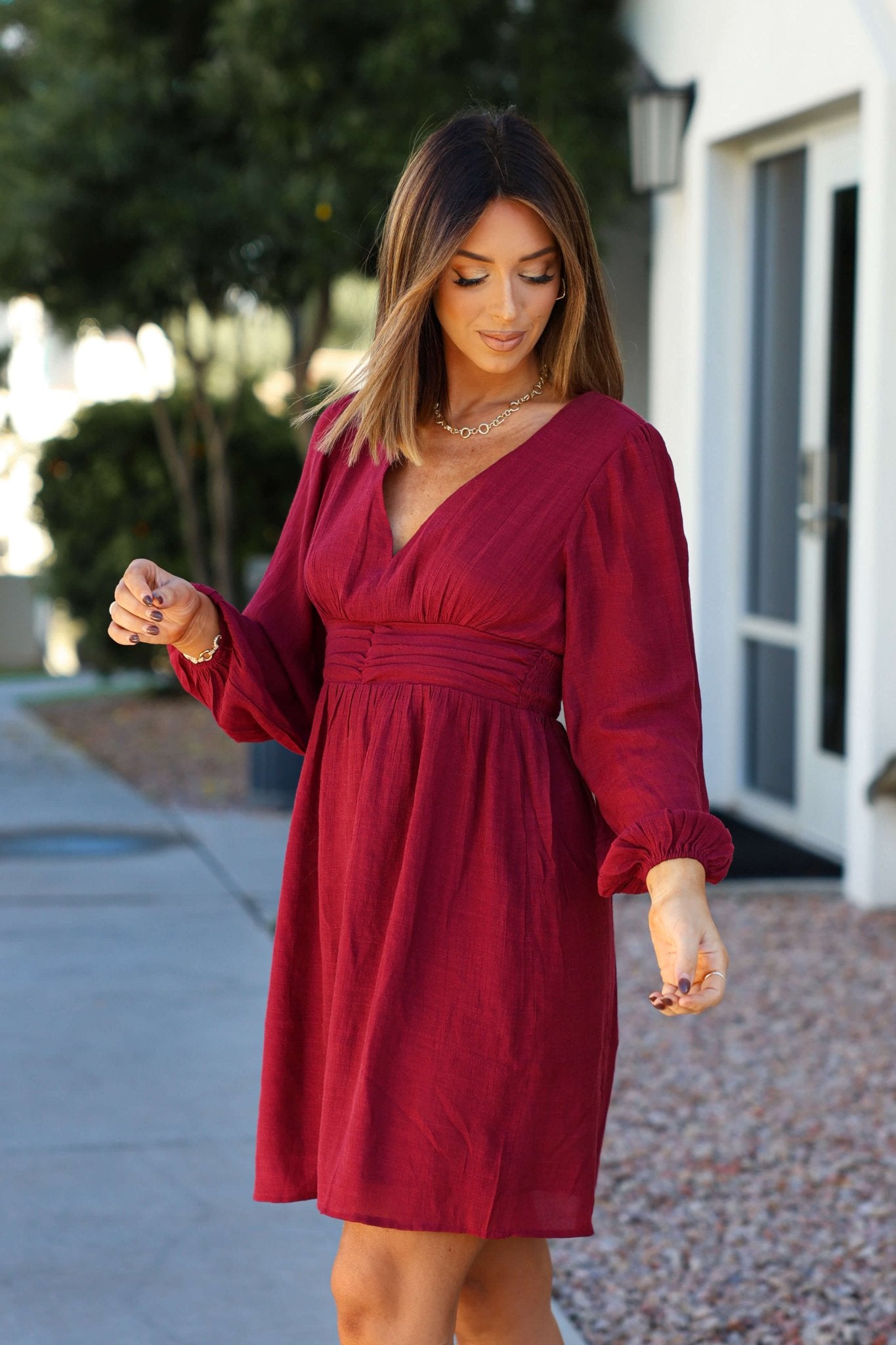 A woman in a Vanessa Burgundy Ruched Mini Dress stands outside a modern building, looking down and gently holding one sleeve.
