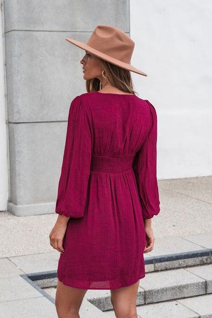 Woman in a Vanessa Burgundy Ruched Mini Dress stands outdoors, facing away toward a light-colored wall.