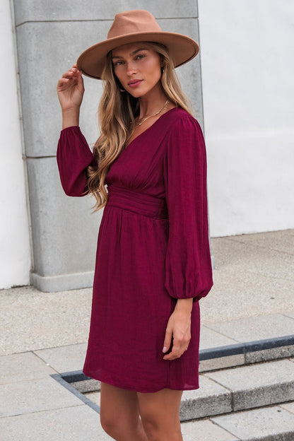 Woman in the Vanessa Burgundy Ruched Mini Dress and tan hat poses outdoors, perfect as a fall wedding guest look.