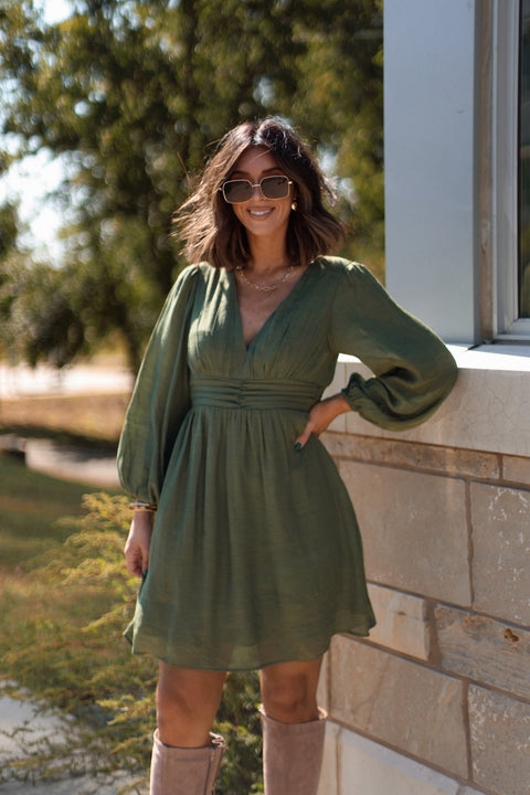 Woman in the Vanessa Green Ruched Mini Dress - FINAL SALE smiles outdoors by a stone wall, wearing sunglasses and hand on hip.